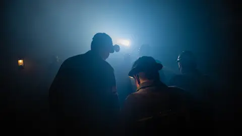 A captivating, almost spiritual, shot of several coal miners at the mouth of a mineshaft. The miners are mostly silhouetted against the soft, ethereal light emanating from within the earth, their features obscured by shadow and coal dust. A few lanterns and headlamps pierce the deep blues and blacks of the mine, highlighting the worn textures of their clothing and equipment. The overall mood is one of mystery and awe, capturing the perilous descent into the earth's depths.