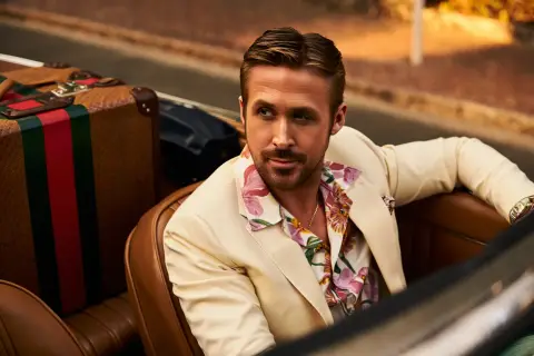 Ryan Gosling, dressed in a sharp cream suit and floral silk shirt, sits behind the wheel of a vintage convertible, the setting sun casting a golden glow on the scene. He is framed by Gucci luggage, the iconic red and green stripes popping against the luxurious leather interior. The image evokes a sense of effortless style and adventure, with a shallow depth of field emphasizing Gosling's charismatic gaze and the allure of the luxury brand.