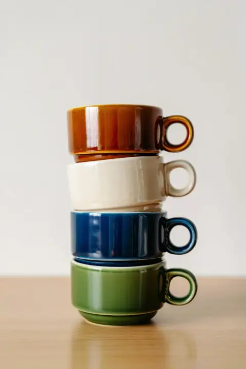 A minimalist product photograph featuring a perfectly balanced stack of four ceramic coffee cups centered against a clean off-white background. The cups are arranged vertically with a brown cup on top, followed by a cream-colored cup, a navy blue cup, and an olive green cup at the bottom providing stability with its small base. Each cup shares the same wide cylindrical design with a circular handle positioned on the right side and features a glossy glazed finish that reflects the even, front-directed artificial lighting. The composition employs a medium shot with shallow depth of field, keeping the cups in sharp focus while subtly blurring the background. The earthy color palette creates a harmonious visual arrangement while showcasing the distinct color of each cup. The photograph's clean, uncluttered aesthetic emphasizes the simple geometric forms and color contrast of the stacked ceramic pieces. Product photography with soft lighting and shallow depth of field against a neutral background.
