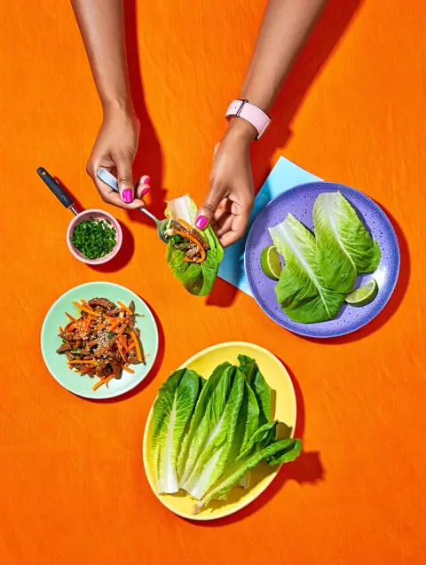An overhead view of a vibrant Asian-inspired lettuce wrap feast arranged on a bright orange textured surface. The composition features multiple colorful plates: a mint green plate with stir-fried meat and shredded carrots sprinkled with sesame seeds in the lower left, a yellow oval plate with fresh romaine lettuce leaves in the bottom center, and a purple-blue speckled plate containing assembled lettuce wraps garnished with lime wedges on the right. A small pink bowl with fresh green herbs sits in the upper left, accompanied by a serving utensil. A pair of medium-toned hands enter the frame from above – the left hand holding a spoon with bright pink nail polish visible, and the right hand displaying a freshly assembled lettuce wrap. The left wrist features a light pink watch. A light blue napkin peeks out from under the purple-blue plate, adding to the colorful arrangement. Overhead food photography with bright direct lighting and defined shadows on a solid colored background.