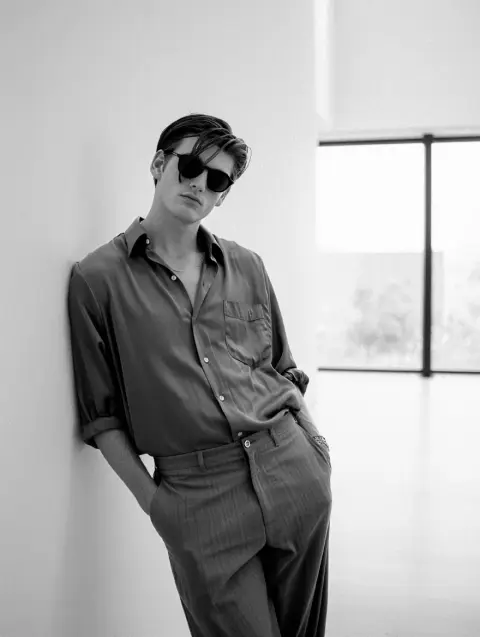 A black and white film photograph from the 1990s shows a man leaning against a wall in a modern art gallery.  He wears loose-fitting clothing, including a gray button-down shirt and dark sunglasses. Natural light enters from a large window behind him, illuminating the minimalist gallery space.  The man's pose is relaxed and contemplative, reflecting the cool aesthetic of the era.