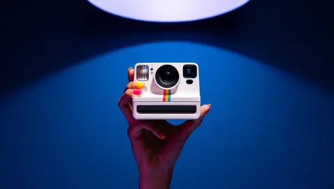 A dramatically lit photograph showcasing a hand with vibrant nail polish holding a white Polaroid Go instant camera against a deep blue background. The camera's rainbow stripe is prominently displayed, catching the light. The image utilizes a single overhead light source, casting soft shadows and creating a sense of depth. The composition remains centered, with a shallow depth of field that emphasizes the camera while slightly blurring the hand. This bold, artistic approach results in a visually striking and contemporary image.